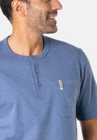Chemise henley bleue en tissu doux, avec un col arrondi, trois boutons et une petite poche poitrine avec une étiquette de marque.