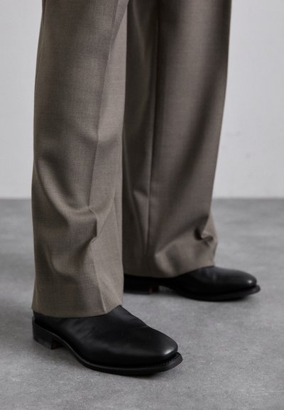 Des chaussures en cuir noir avec un design à bout pointu, associées à un pantalon gris sur mesure. Les chaussures affichent un talon bas et une texture lisse.