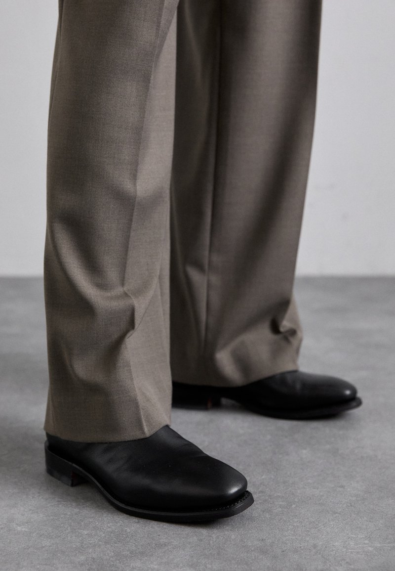 Black leather shoes with a pointed toe design paired with gray, tailored trousers. Shoes feature a low heel and smooth texture.
