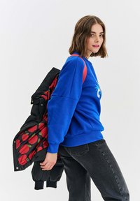 Mladá žena ve modré mikině a černých džínách nese přes rameno černou prošívanou bundu s podšívkou v červené jablečné vzoru.