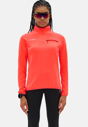 Persoon met gevlochten haar, draagt een rood langemouw-trui met rits, zwarte legging, zwarte smartwatch en reflecterende sportzonnebril, staand tegen een effen achtergrond.