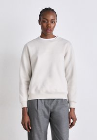 Lys grå sweatshirt med rund halsutskjæring og lange ermer, laget av mykt stoff. Har en subtil logo detaljer på forsiden.