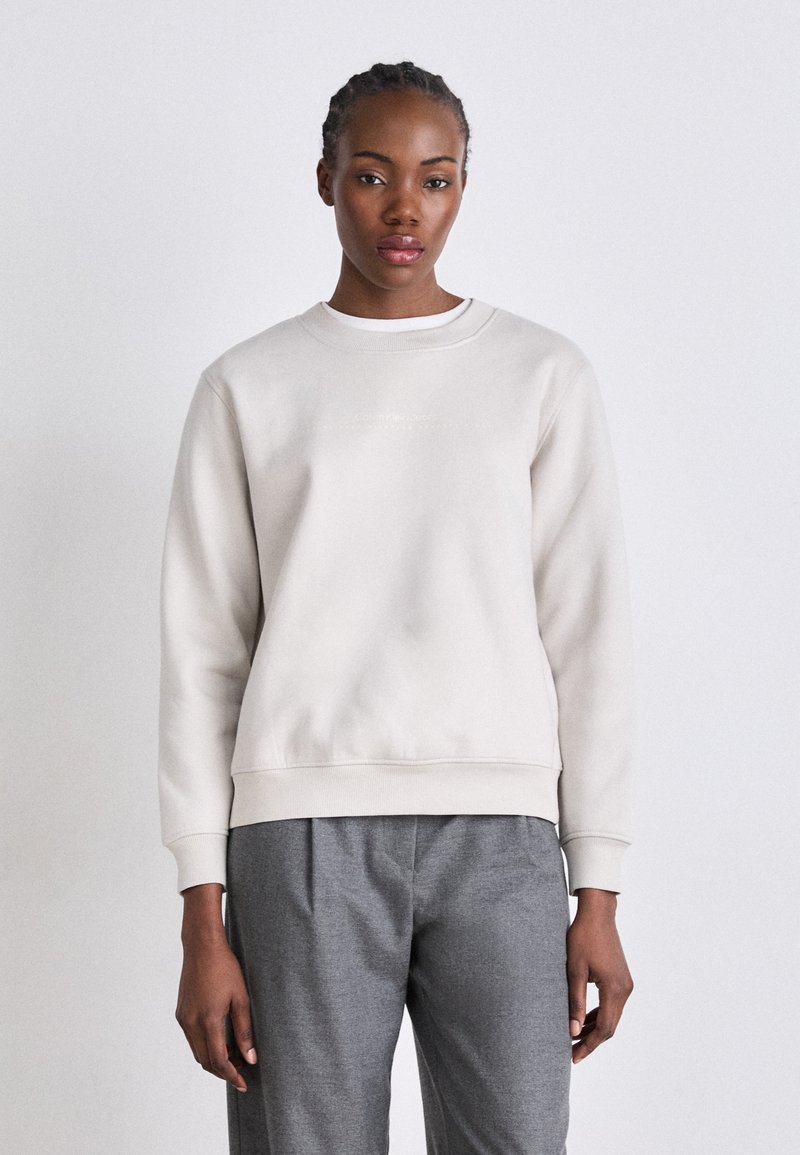 Lys grå sweatshirt med rund halsutskjæring og lange ermer, laget av mykt stoff. Har en subtil logo detaljer på forsiden.