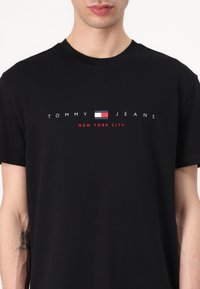 Schwarzes Baumwoll-T-Shirt mit dem Schriftzug "TOMMY JEANS" und einem kleinen rot-weiß-blauen Logo. Rundhalsausschnitt und kurze Ärmel.