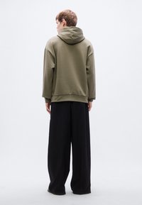 Φούτερ με κουκούλα σε ελαιώδες πράσινο, με χαλαρή εφαρμογή και χαμηλωμένους ώμους, συνδυασμένο με άνετο μαύρο παντελόνι. Λείο ύφασμα, με ελάχιστες λεπτομέρειες.