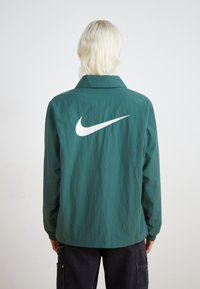 Zöld, könnyű Nike dzseki, fehér logóval a hátán, klasszikus gallérral és rugalmas mandzsettákkal a laza illeszkedés érdekében.