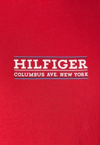 Tessuto rosso con testo bianco che recita "HILFIGER" e "COLUMBUS AVE. NEW YORK." Presenta un design semplice con caratteri in grassetto.