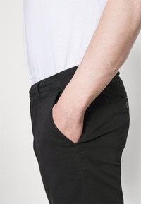 Zwarte broek met een gladde textuur, voorzien van zijzakken en een aansluitend ontwerp. Draagt met een wit shirt, wat een ontspannen uitstraling laat zien.