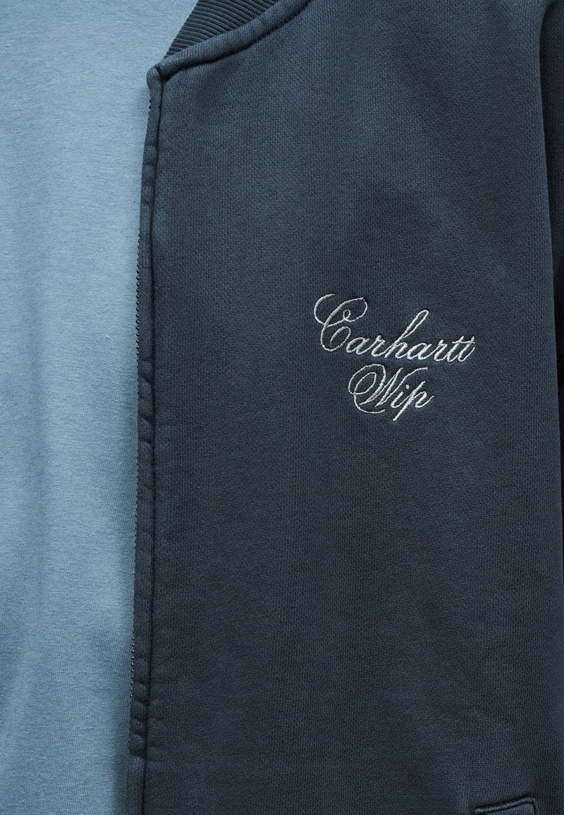 Close-up of a blue and navy Carhartt Wip sweatshirt with embroidered white logo on the chest and visible zipper seam.