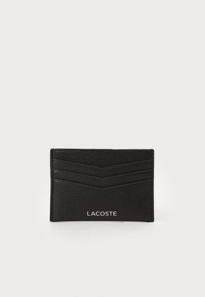 Porte-cartes en cuir noir avec une finition texturée, comportant trois fentes pour cartes, des rayures diagonales, et un logo Lacoste blanc en bas.