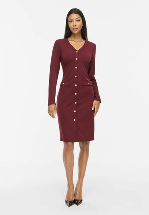 Modèle portant une robe bordeaux au genou, boutonnée sur le devant, avec un col en V et des rabats de poches, associée à des escarpins noirs sur un fond blanc uni.