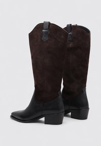 Botas de ante marrón hasta la rodilla con punta negra texturizada y tacón de bloque, que presentan detalles de cuero negro en la parte superior.