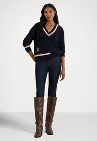 Laivastonsininen palmikkoneulepaita V-pääntiellä, jossa viininpunaiset ja kermaiset raidat helmassa ja kalvosimissa sekä kirjailtu logo; yhdistetty tummiin farkkuihin ja ruskeisiin polvipitkiin saappaisiin.