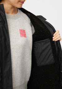 Giacca nera esterna con una fodera interna morbida e testurizzata. La felpa grigia sottostante presenta un piccolo design grafico rosso. La giacca ha una tasca.