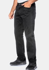 Czarne bawełniane spodnie o dopasowanym kroju, z zamkiem błyskawicznym, zapięciem na guzik, pięcioma kieszeniami i kontrastowymi szwami. Noszone z czarnymi butami.