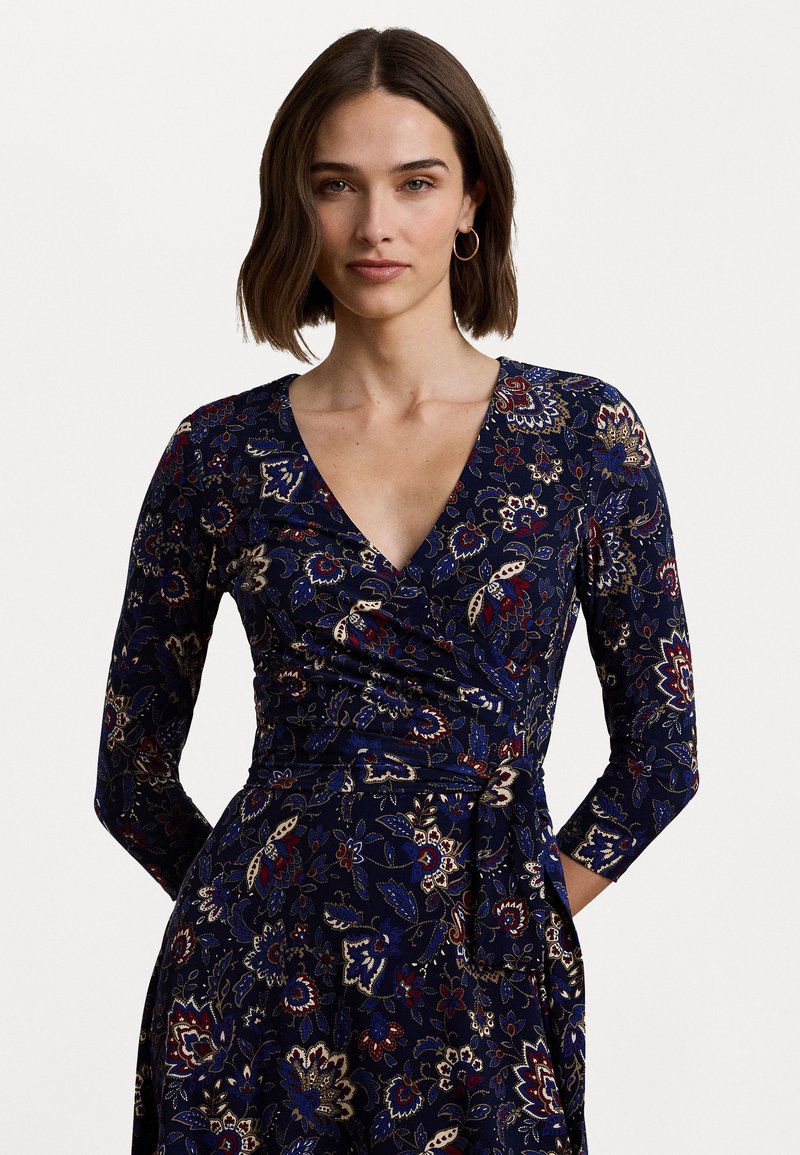Abito avvolgente blu navy con maniche lunghe, caratterizzato da un motivo floreale in oro, rosso e blu. Tessuto morbido e testurizzato con scollatura a V e dettaglio della vita annodata.