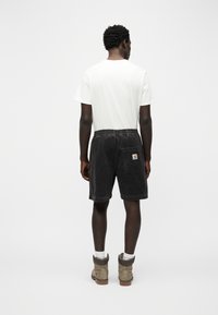 Άνδρας που κοιτάζει προς τα πίσω, φοράει λευκό t-shirt, μαύρα cargo σορτς με λογότυπο στην πίσω τσέπη, λευκές κάλτσες και μπεζ μπότες εργασίας σε απλό φόντο.