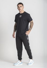 Svart bomulls t-shirt med en vit logotyp på bröstet, tillsammans med svarta joggers med vita mönster. Skorna är blå och vita sneakers.