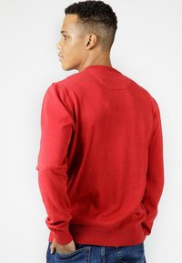 Czerwona swetrowa bluzka z długimi rękawami, ściągaczami przy nadgarstkach i u dołu, gładka tekstura, design z okrągłym dekoltem, noszona z niebieskimi dżinsami.