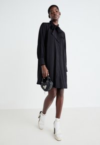 Robe noire à manches longues avec un col à nouer, en tissu doux, coupe décontractée, longueur genoux. Se marie bien avec un sac à main noir en forme de cœur et des talons blancs à lanières.