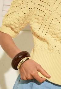 Suéter amarillo de punto con patrón texturizado y puños acanalados, combinado con una pulsera marrón brillante y una pulsera de acento metálico dorado.