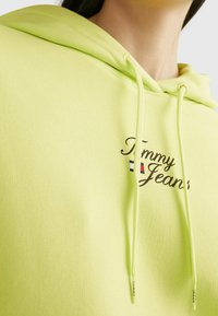 Neongrön hoodie med snörning, med broderad "Tommy Jeans"-logotyp och en liten tri-färg accent (blå, vit, röd) på bröstet.