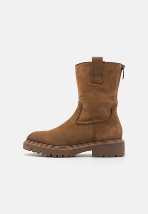 Brown suede ankle boot with a rounded toe, side zipper, and a pull tab. Features a textured rubber sole. Casual and robust design.