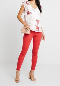Blusa bianca con motivo floreale rosso, scollatura a V, maniche a campana; jeans skinny rossi; borsa a tracolla rosa; sandali con tacco quadrato color nudo.