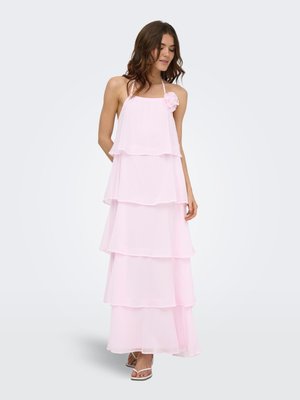 Vrouw draagt een lichtroze gelaagde halter maxi-jurk met een bloemdetail nabij de halslijn, staand op een witte achtergrond.
