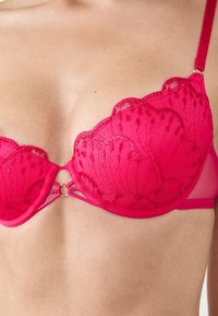 Sujetador acolchado en fucsia con detalles de encaje, bordes festoneados y un pequeño acento circular dorado en el centro. Tirantes ajustables y tejido transparente.