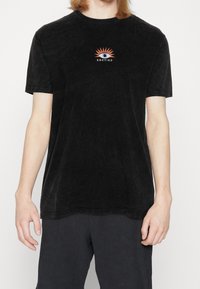 Camiseta negra de manga corta hecha de tejido suave, con un gráfico colorido de un sol y un ojo, y el texto "KAOTIKO" debajo. Cuello redondo.