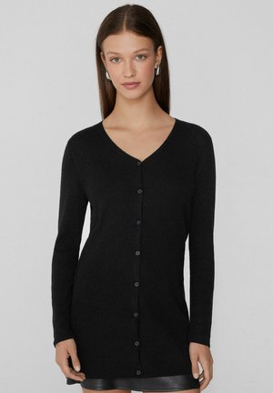 Mladá žena se rovnými hnědými vlasy nosí dlouhý černý kardigan na knoflíky přes černou sukni, stojí na pozadí s jednoduchým světlým pozadím.