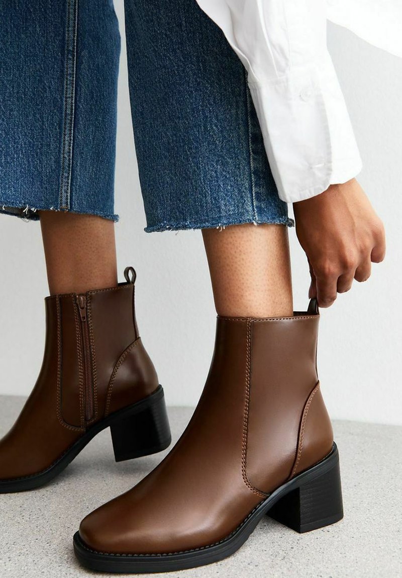 Bottines en cuir marron avec un orteil arrondi, une fermeture éclair sur le côté et un talon carré, associées à un jean en denim déchiré et cropped.