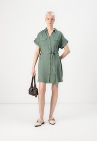 Abito camicia verde a maniche corte con bottoni anteriori, due tasche sul petto, vita legata e tessuto testurizzato. Abbinato a scarpe beige e una borsa marrone.