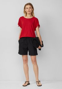 Rode top met korte mouwen en strikdetails, gecombineerd met zwarte opgerolde shorts. Het model houdt een zwarte gehaakte tas vast en draagt platte sandalen.