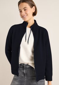 Marinblå vadderad jacka med hög krage och framsida dragkedja, med dragsnören och en texturerad yta, bärs över en ljus tröja.