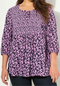 Blusa floreale rosa con motivi neri, corpetto smockato e maniche a tre quarti. Presenta un collo arrotondato con un dettaglio a fiocco.