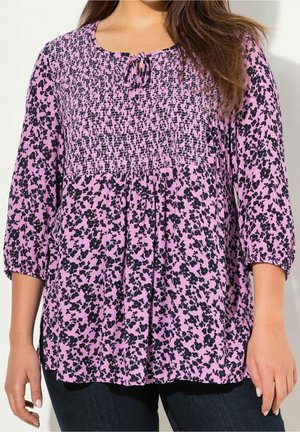 Blouse florale rose avec des motifs noirs, corsage smocké et manches trois-quarts. Dotée d'un col rond avec un détail à nouer.