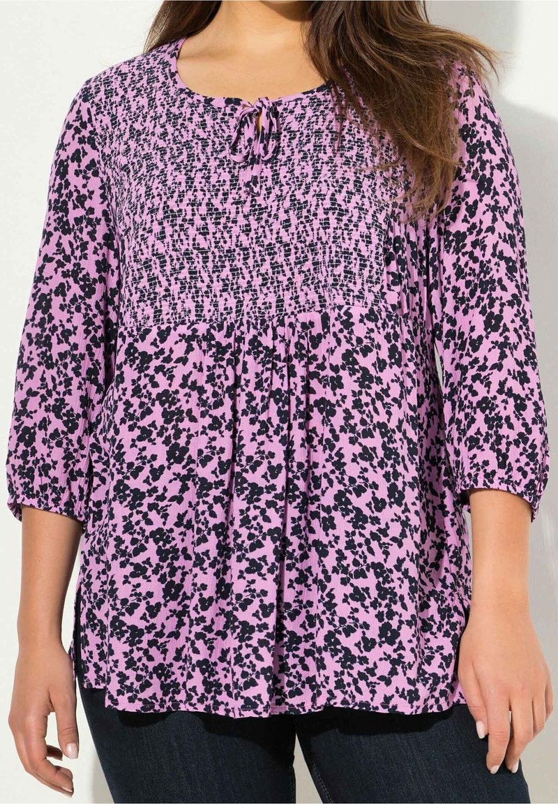 Blusa floreale rosa con motivi neri, corpetto smockato e maniche a tre quarti. Presenta un collo arrotondato con un dettaglio a fiocco.