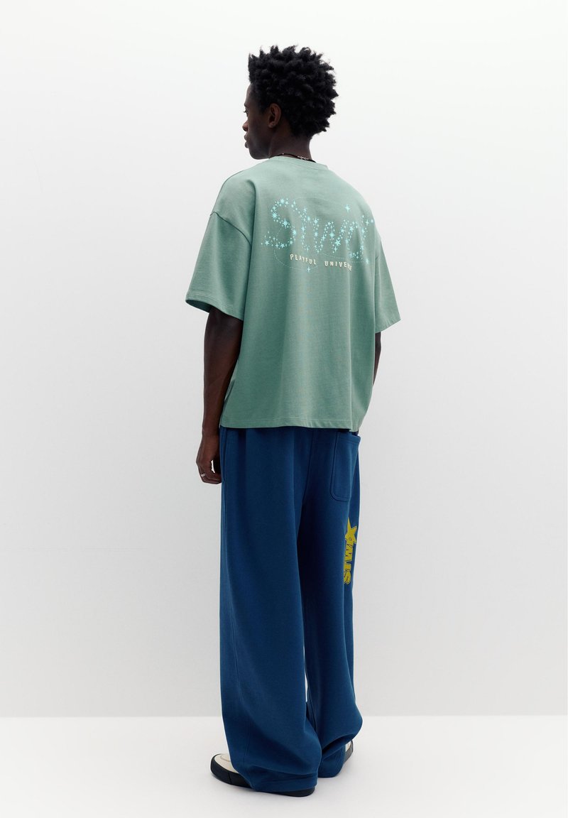 Persoană cu păr scurt și creț purtând un tricou verde larg cu design stelar și pantaloni largi albaștri cu inscripție galbenă, stând cu fața spre stânga.