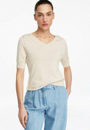 Kobieta z włosami zaczesanymi do tyłu, ubrana w beżową bluzkę z krótkim rękawem i dekoltem w serek oraz luźne, jasnoniebieskie dżinsy, stojąca z jedną ręką w kieszeni.