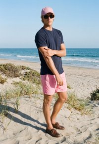 Junger Mann mit pinker Kappe, schwarzer Sonnenbrille, marineblauem Hemd und pinken Shorts, der am sandigen Strand nahe dem Ozean mit spärlicher Dünenvegetation steht.
