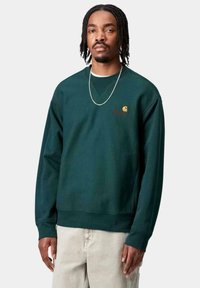 Sudadera verde oscuro de mangas largas, con cuello redondo y puños acanalados. Presenta un pequeño logo naranja en el pecho. Textura de tela suave.