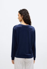 Γυναίκα με μακριά σγουρά σκούρα μαλλιά που φοράει μπλε σκούρο μακρυμάνικο τοπ και ανοιχτό γκρι παντελόνι, γυρισμένη με την πλάτη σε λευκό απλό φόντο.