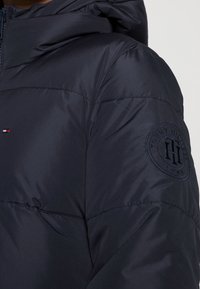 Námořnická puffer bunda s kapucí, s prošívanou texturou. Na levém rukávu je vyšívané kruhové logo se značkou "Tommy Hilfiger".