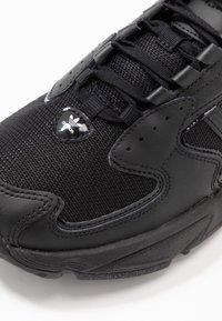 Svart atletisk sko med en ovandel av mesh och läder, texturerad yta, tonala accenter och en glansig logotypdetalj på snörningsområdet.