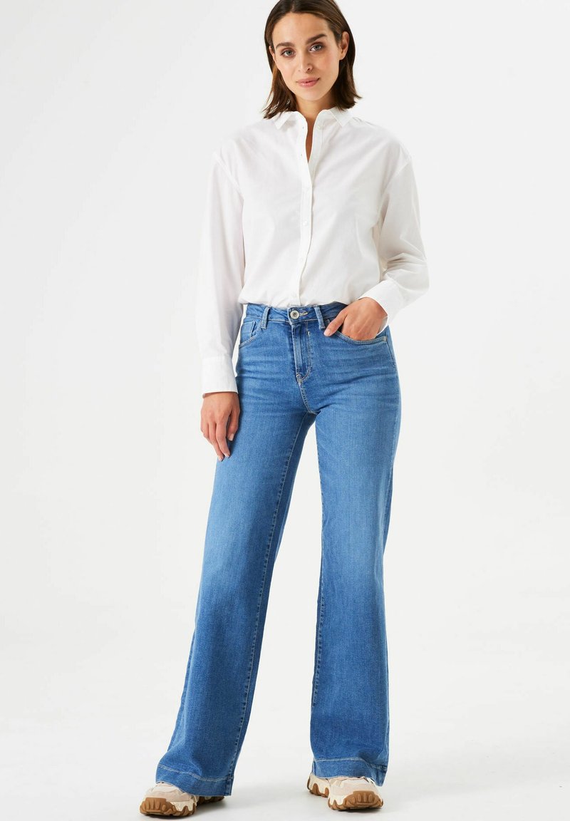 Frau steht mit einer Hand in der Tasche, trägt ein weißes Hemd mit Knöpfen, blaue weite Jeans und beige Sneakers vor einem schlichten weißen Hintergrund.