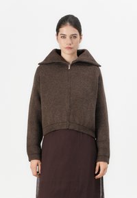 Brun ullblandingsjakke med høy krage, glidelås foran og ribbestrikket nederkant. Kuttet design med lange ermer. Kombinert med en mørk kjole.