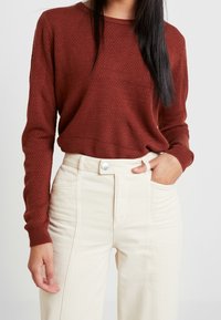 Bordeaux kleurige gebreide trui met lange mouwen en ronde hals; gecombineerd met high-waisted off-white broek met een sluiting aan de voorkant.