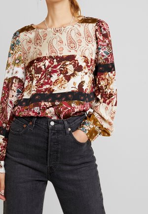 Femme portant un chemisier patchwork à manches longues avec des motifs paisley et floraux rentré dans un jean noir taille haute, main dans la poche.